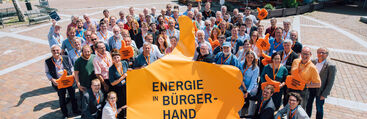 Viele Menschen als Gruppe draussen mit großem orangem Banner in der Hand und dem Slogan "Energie in Bürgerhand"