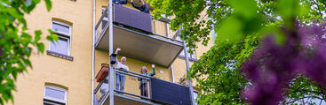 Winkende Menschen von drei Balkonen an denen ein Solarmodul/Balkonmodul befestigt ist